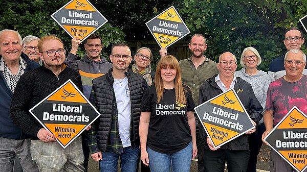 An enthusiastic group of Liberal Democrat members and supporters are gathered on the stone steps of Grimsby Town Hall, holding bright yellow ‘Liberal Democrats Working For You’ signs and raising their arms in a show of unity. They stand ready for the upcoming elections. At the front and centre is Cllr Nicola Aisthorpe, North East Lincolnshire Lib Dem Group Leader, wearing a light-coloured coat over a black dress and smiling confidently.