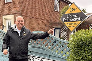 Brian Barrett standing outside a home on Barton Grove, pointing to a Liberal Democrat “Working For You” garden poster.